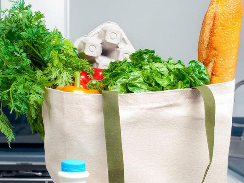 Carrots, eggs, peppers, leafy greens and a loaf of french bred in a cloth grocery bag.