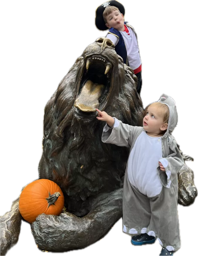 Two young children in Halloween costumes interacting with a lion sculpture; one child dressed as a pirate standing on the sculpture, and the other, in a mouse costume, touching the lion's tongue.