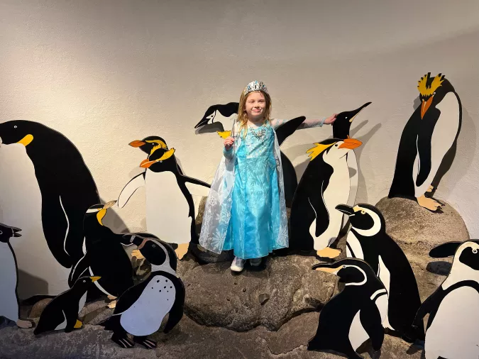 A young girl dressed as a princess with a tiara, posing with painted penguin cutouts at a zoo exhibit.