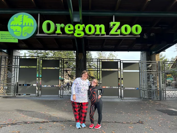 Two people posing happily in front of the Oregon Zoo entrance, one in a festive pajama outfit and the other in a sporty black and red tracksuit.