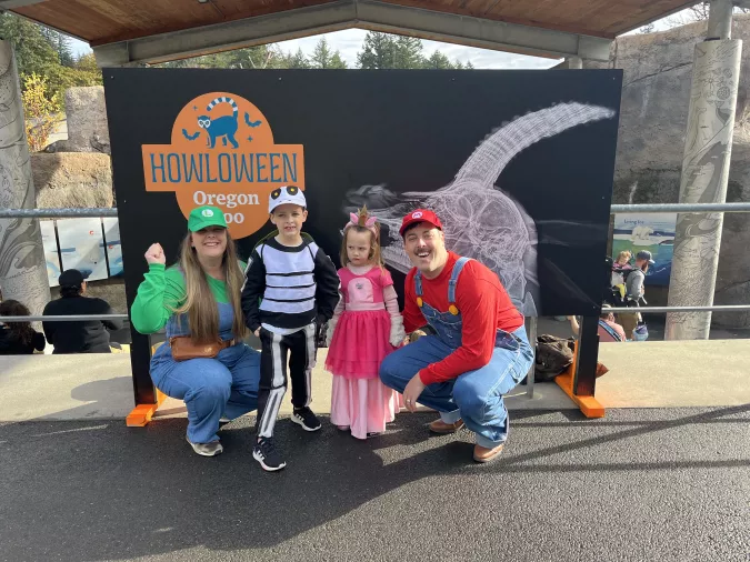 Family of four in Halloween costumes posing at an Oregon Zoo event; includes a woman dressed as Luigi, a man as Mario, a boy as a skeleton, and a girl in a princess costume, all smiling in front of a 'Howloween Oregon Zoo' sign.