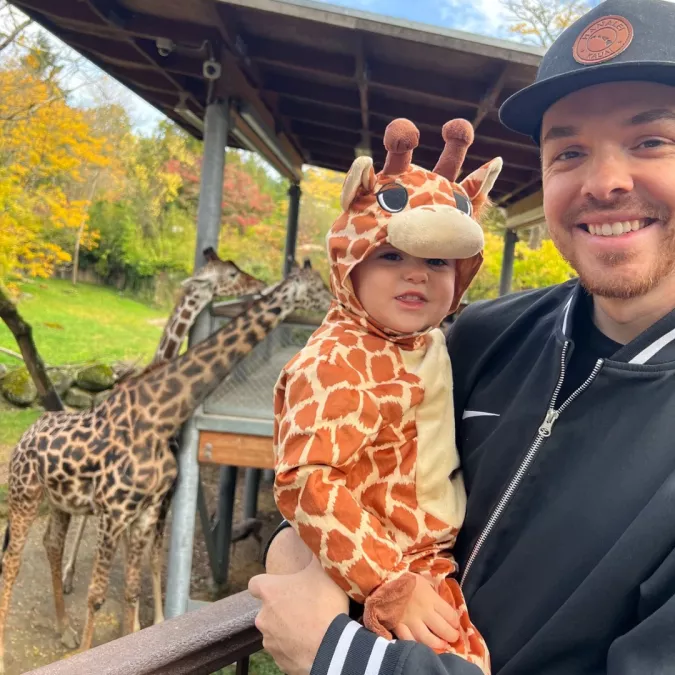 A smiling man holds an infant dressed as a giraffe in his arms, 2 giraffes are visible in the background