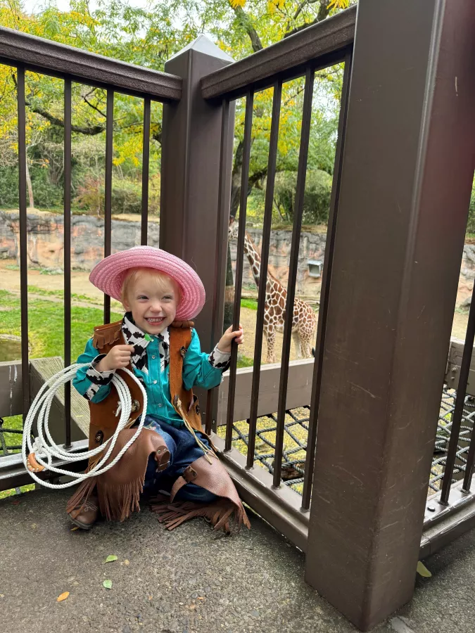 Young child dressed as a cowgirl, complete with a hat, fringed vest, and chaps, happily posing with a lasso near a giraffe exhibit at a zoo.