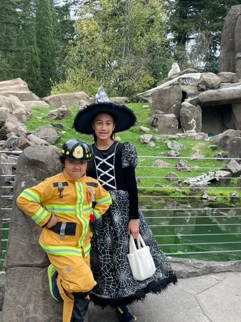 Two children in Halloween costumes at the zoo: one dressed as a firefighter and the other as a witch, posing in front of a rocky exhibit with a white mountain goat visible in the background.