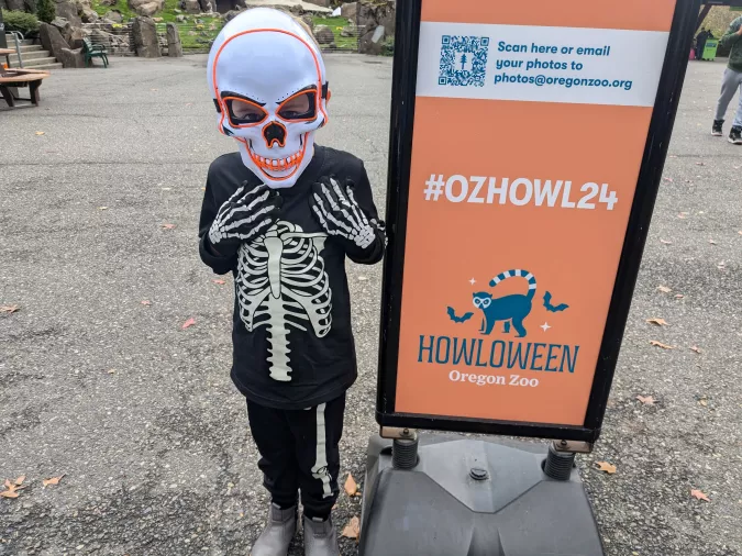 Child in a skeleton costume with a skull mask standing next to a Howloween event sign at the Oregon Zoo. The sign includes the hashtag #OZHOWL24 and a QR code for sharing photos.