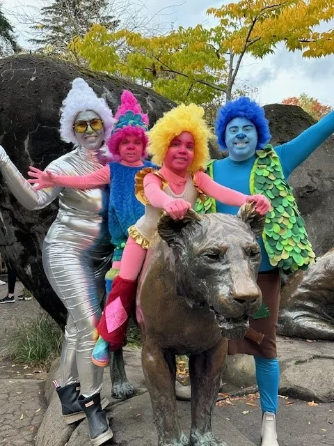 Group of people dressed in colorful, whimsical costumes with vibrant wigs, standing around and sitting on a statue of a large cat at the zoo. One person is in a silver metallic outfit with a white wig, while others wear bright blue, yellow, and pink wigs and matching costumes, creating a fun and festive atmosphere.