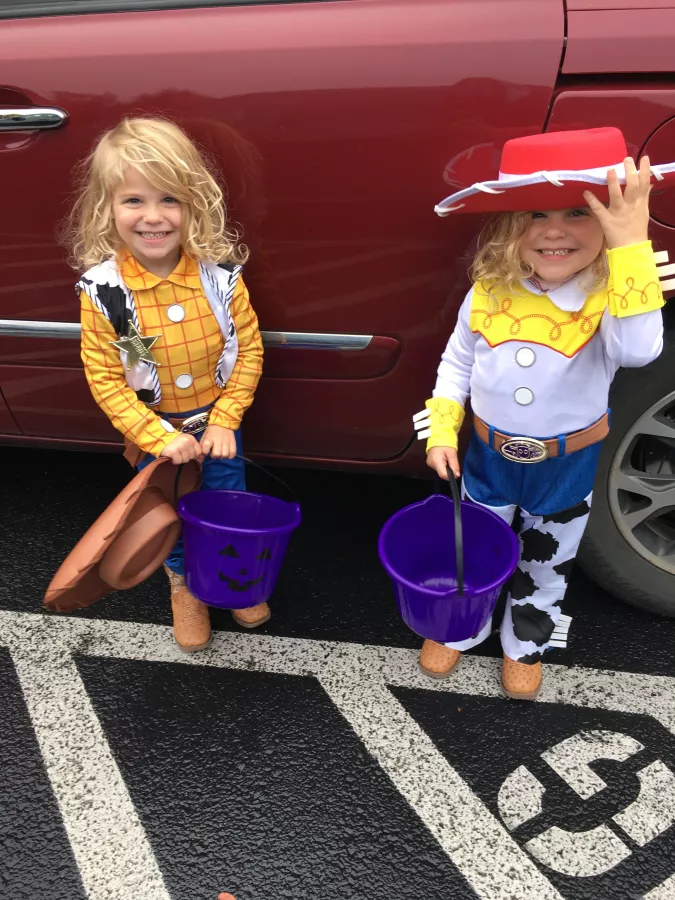 Two young children dressed in cowboy-themed costumes, resembling Woody and Jessie from Toy Story. They are holding purple trick-or-treat buckets and standing in front of a red car, smiling and ready for Halloween festivities.