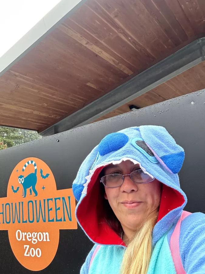 Person in a blue costume hoodie with large eyes on the hood, standing in front of a 'Howloween Oregon Zoo' sign. The person is wearing glasses and has a slight smile, ready to celebrate the Halloween-themed event at the zoo.