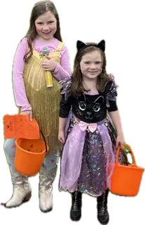 Two young girls in Halloween costumes holding orange trick-or-treat buckets. One is dressed as a singer with a gold-fringed outfit, a pink shirt, white boots, and holding a toy microphone. The other is dressed as a sparkly black cat with cat ears, wearing a purple skirt and black boots. Both are smiling and ready for Halloween festivities.