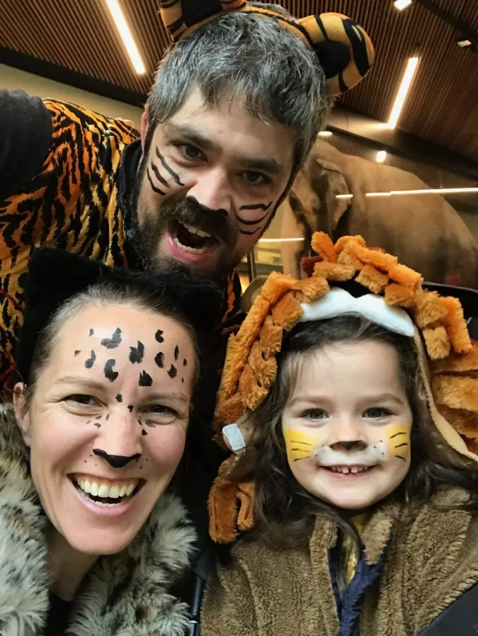 Family dressed in animal-themed costumes with face paint, smiling and posing for a photo. The adults have tiger and leopard face paint, while the child is dressed as a lion with face paint and a mane hood. They appear to be enjoying a fun day at an animal-themed event.